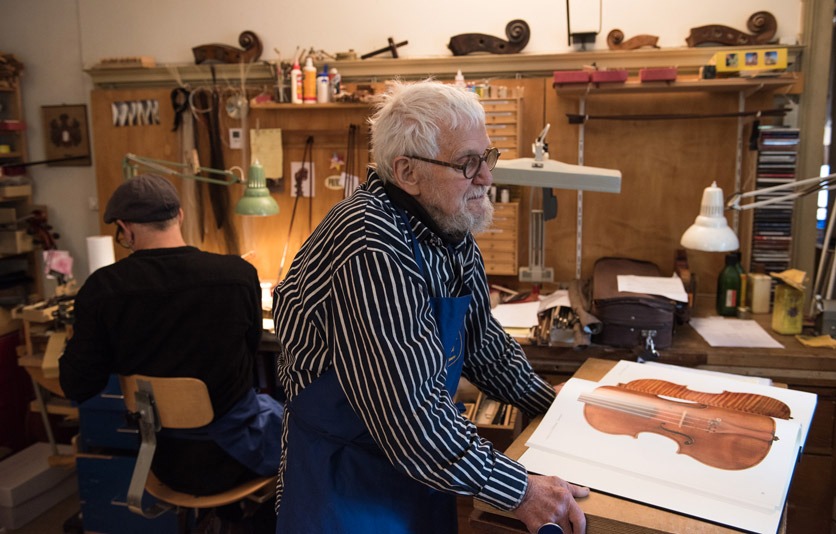 Geigenbauer Rast mit seinem Sohn im Atelier. Sein Sohn arbeitet am Schreibtisch, während er Unterlagen studiert. 