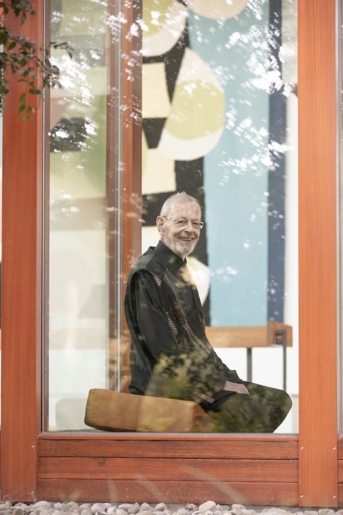 Portrait von Niklaus Branschen, Zen-Meister, im Meditationssitz hinter einer Fensterscheibe, lächelnd.