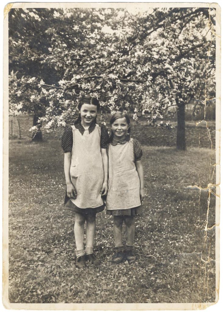 Schwarzweissfoto: Zwei Mädchen mit Schürzen halten sich an der Hand.