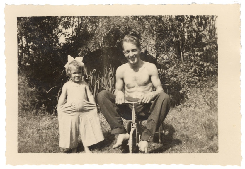 Anno 1943: Edith Baumgartner und ihr Vater im Garten. Sie trägt eine Schleife im Haar. Der Vater sitzt mit freiem Oberkörper auf einem Kinderfahrrad.