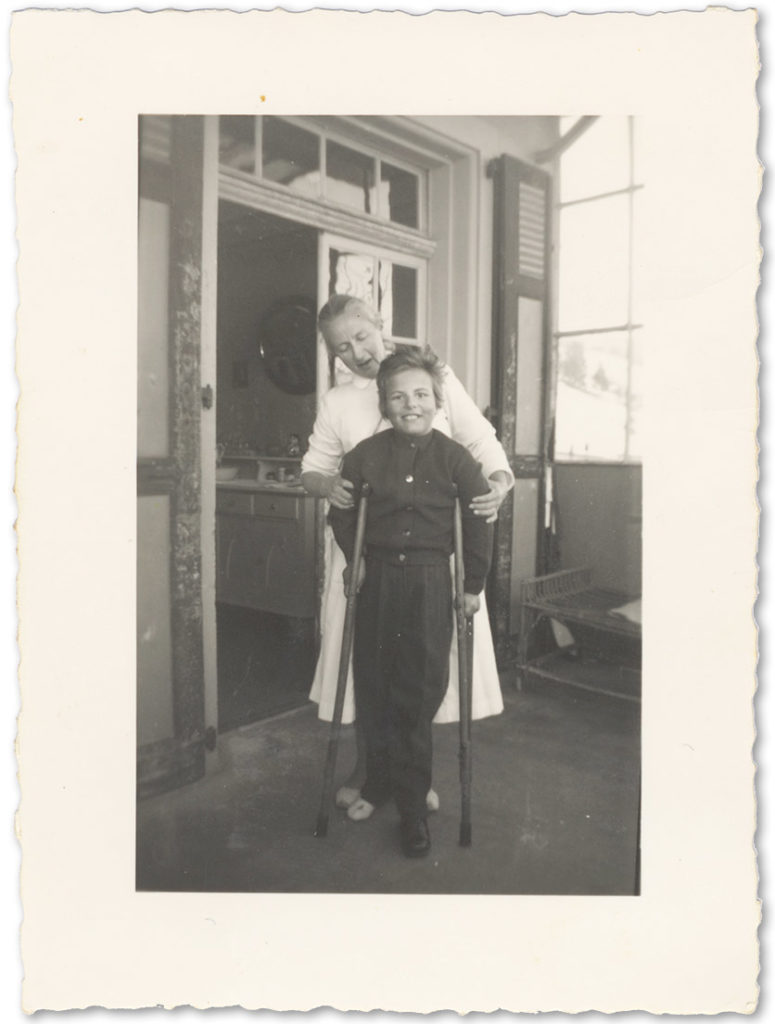 Schwarzweissfoto von 1958: ein kleines Mädchen mit Krücken, dahinter steht eine Krankenschwester in Uniform. 