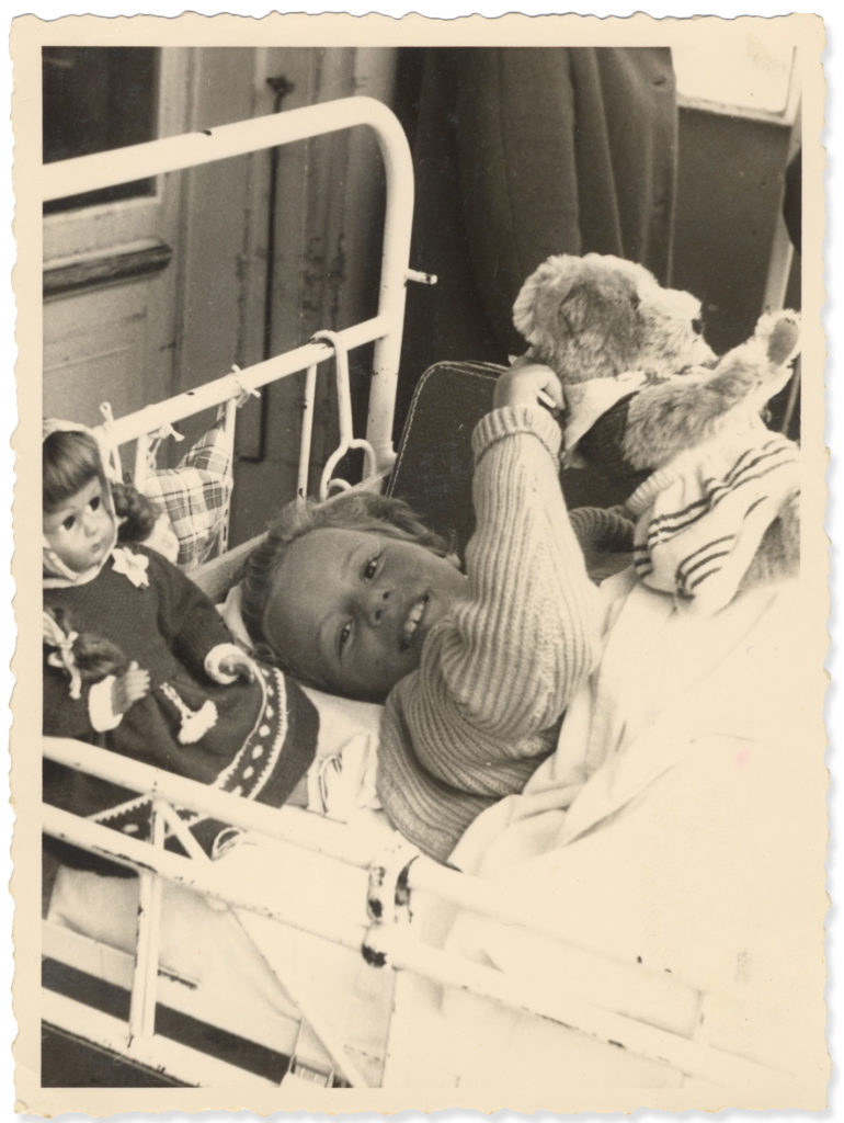 Schwarzweissbild von 1958: ein Mädchen liegt in einem weissen Gitterbett, lächelt in die Kamera und hält einen Teddybären.