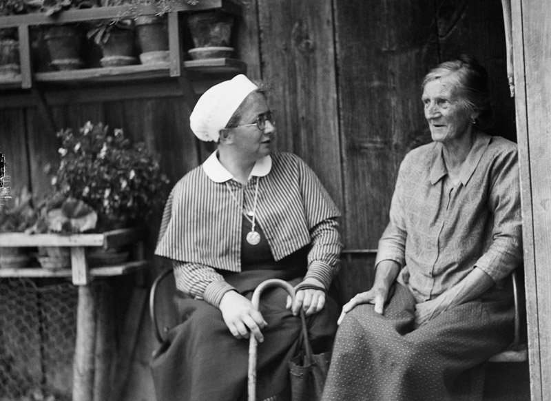 Fürsorge um 1923: eine Gemeindeschwester sitzt neben einer alten Frau vor dem Haus.