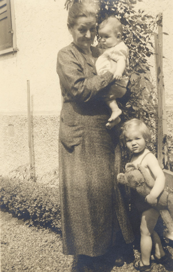 Im Garten vor dem Haus: Die Grossmutter von Maya Lütolf-Busch aus Baar ZG, im Hauskleid, hat ein Enkelkind auf dem Arm, eines steht mit Teddy neben ihr.