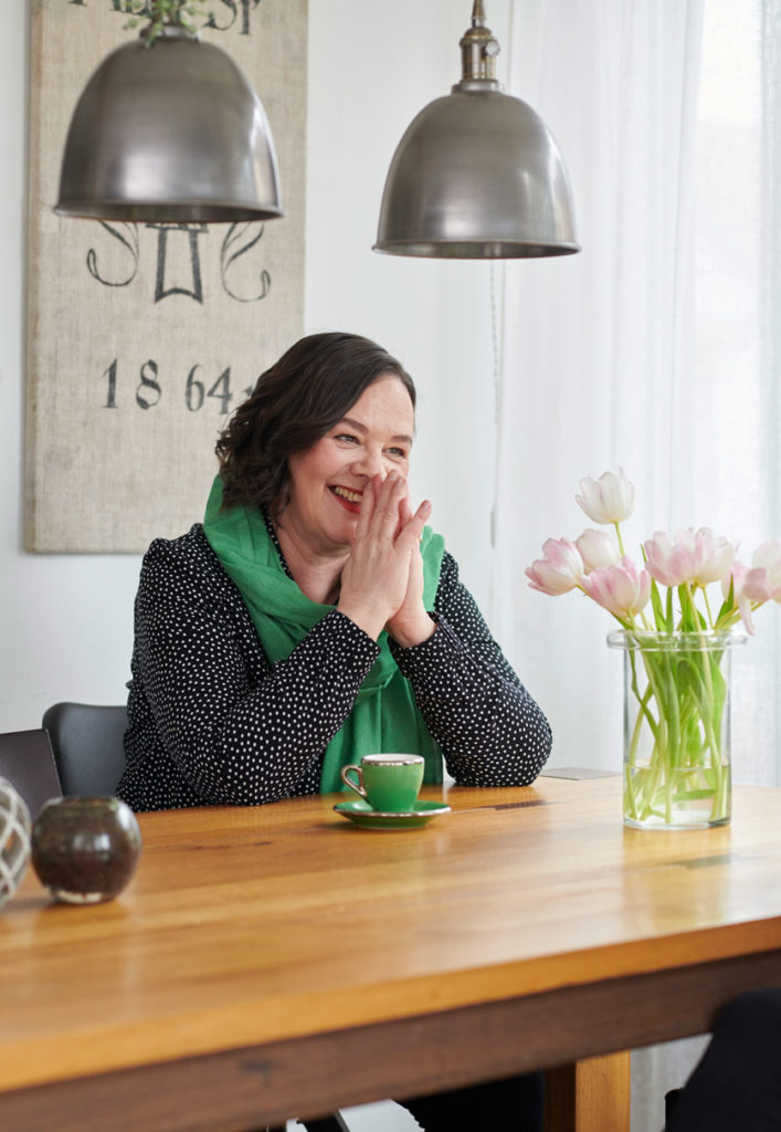 Interviewbild mit Vera Kaa. Sie sitzt in ihrer Wohnung an einem Holztisch, darauf eine Vase mit rosa Tulpen. Sie lächelt und hält die gefalteten Hände vors Gesicht.