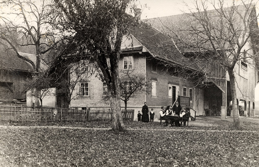 Ca. 1918 im Aargauer Freiamt. Man sieht einen grossen Bauernhof. Davor sitzen ein paar Männer in Sonntagskleidern an einem TIsch.