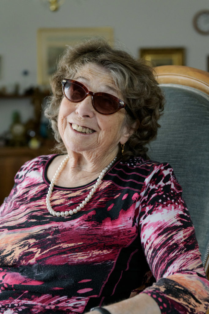 Portrait von Cornelia Zumsteg in ihrem Sessel. Sie trägt eine Sonnenbrille, eine weisse Perlenkette und eine violett-gemusterte Bluse. Sie lächelt in die Kamera. Die blinde Cornelia Zumsteg hört sich gerne die Zeitlupe an, bereitgestellt von der Blindenbibliothek.