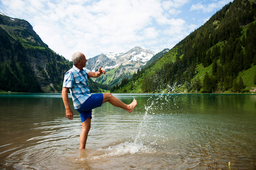 Alleinsein als Chance: Ein Senior steht an einem Bergsee und spritz mit den Füssen Wasser umher.