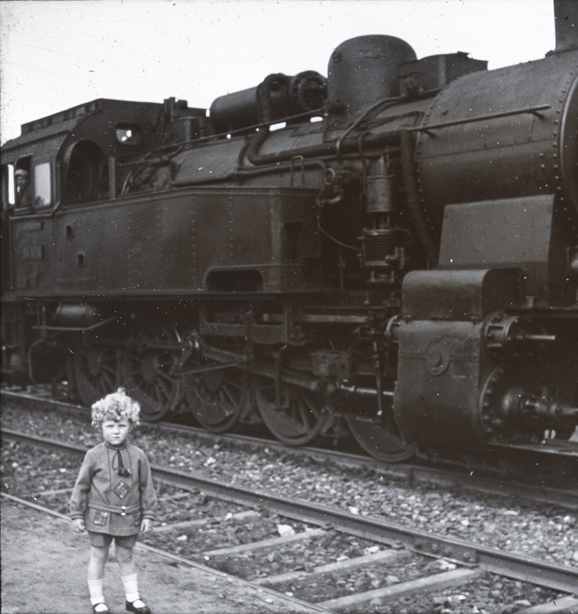 Anno 1930: Ein kleiner Junge mit blonden Locken steht mit ernstem Blick vor einer Dampflok. 