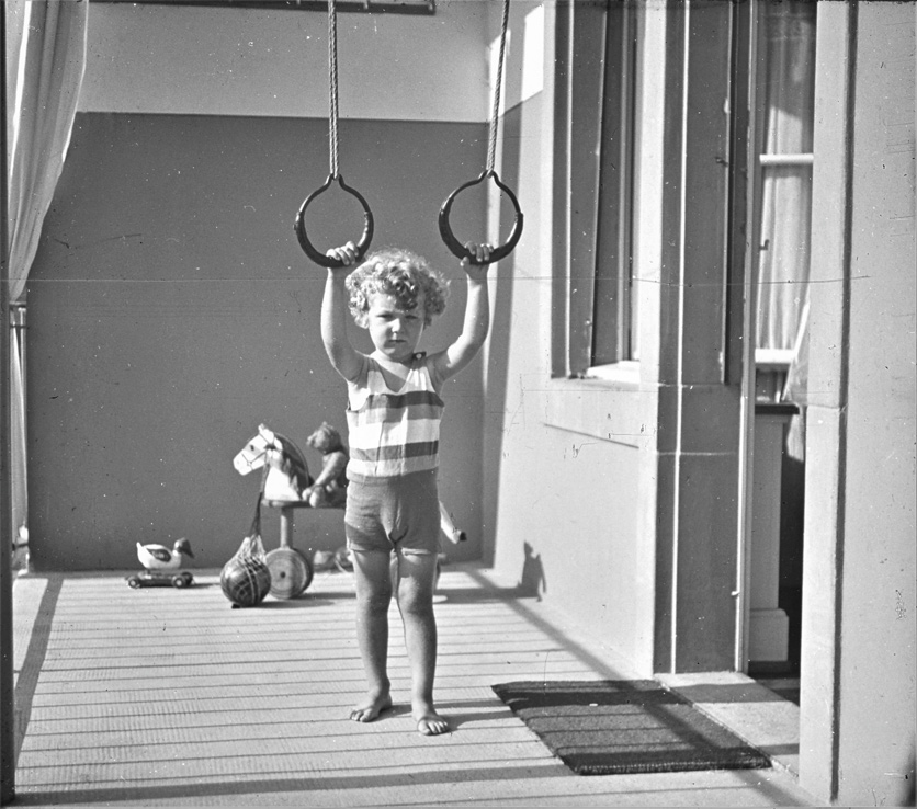 Anno 1930: Ein Junge mit blonden Locken spielt auf einer Terrasse und hält sich Turnringen, die von der Decke hängen. Im Hintergrund sieht man einen Ball und ein Holzpferd auf Rädern. 