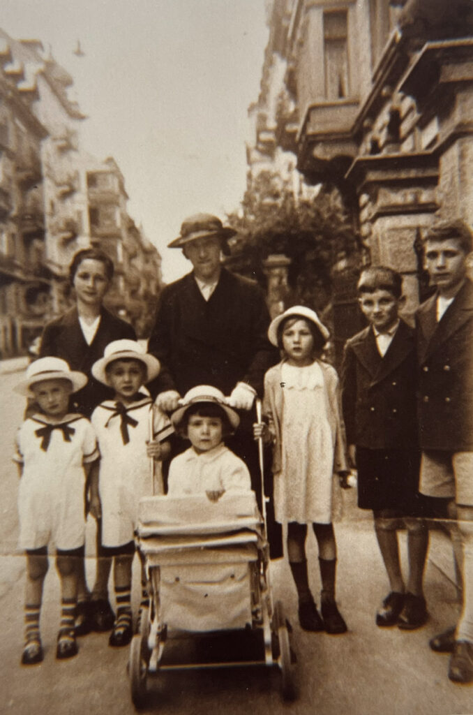 Anno 1935: Familienfotomit Béatrice Romano im Kinderwagen. Die Kinder sind im Sonntagskostüm, vor städtischem Hintergrund.