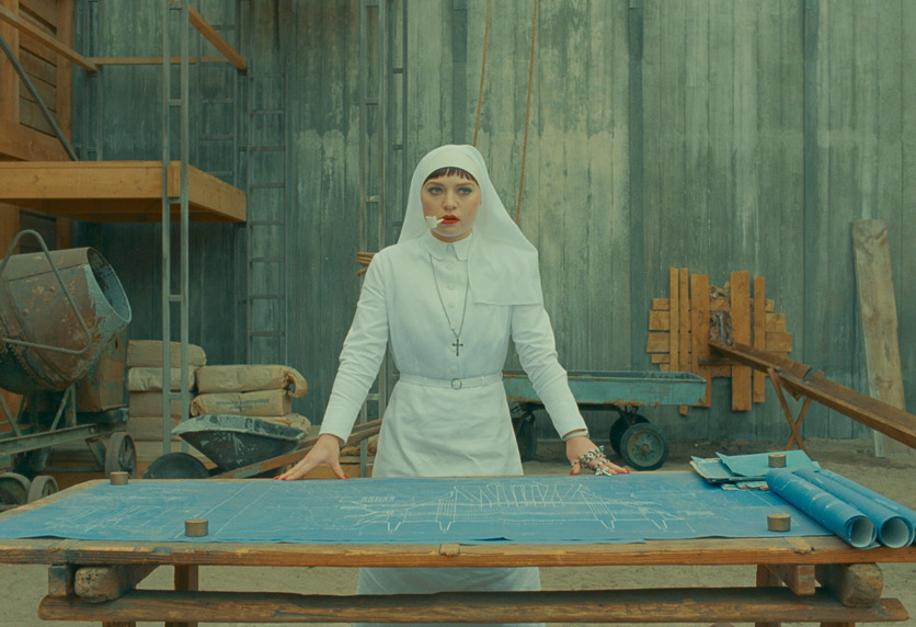 Mia Threapleton als Tochter Liesl, eine Nonne. In einem weissen Kostüm steht sie vor Bauplänen und raucht Pfeiffe.
